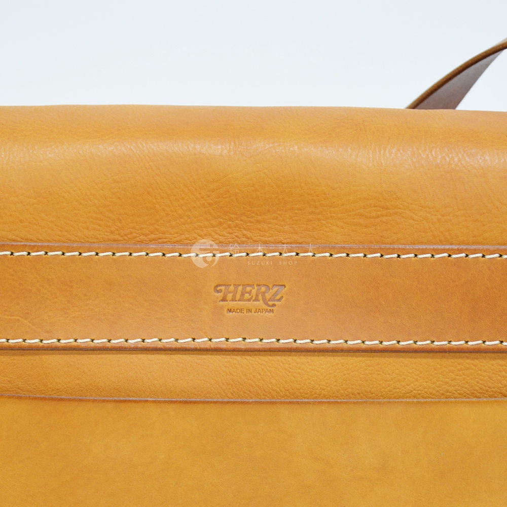 職人手作皮革復古翻蓋郵差包，採用淺棕色皮革，表面有細緻紋理，配有白色縫線裝飾，中央皮革壓印有「HERZ」品牌標誌及「MADE IN JAPAN」字樣。