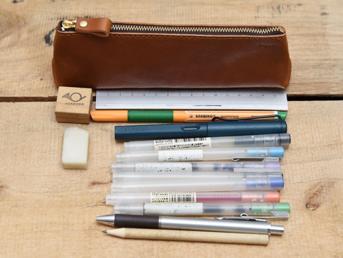 職人手作皮革Organ三角收納筆袋 (GS-28) M，咖啡色皮革材質，配有金色拉鍊。筆袋旁有橡皮擦、木質印章、金屬尺、螢光筆、鋼筆及多支透明筆管的筆。