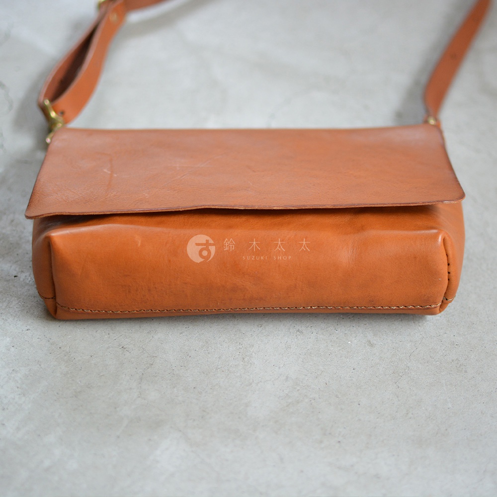 一個棕褐色的皮革斜背包，有著簡約的翻蓋設計，背包的皮革表面帶有自然的紋理，下方印有SUZUKI SHOP的標誌。