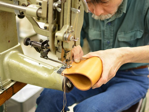 一位工匠正使用工業縫紉機縫製一個淺棕色的皮革筆筒。皮革製品的邊緣有縫線，顯示出精緻的工藝。