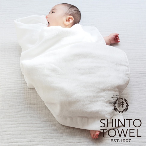 一件白色的2.5層紗嬰兒浴巾，柔軟親膚，包裹著一個嬰兒，浴巾上有品牌標籤。