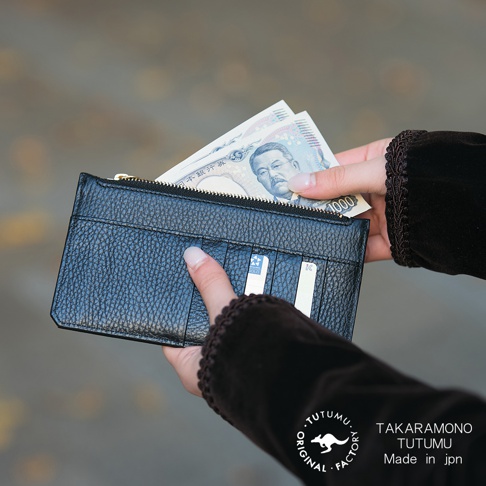 一手拿著黑色皮革長夾，夾中有日圓鈔票，另一手放在夾子側面，鈔票面額為1000日圓，左下方有TUTUMU的標誌和文字TAK AR A MONO TUTUMU Made in jpn。