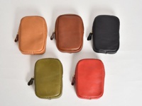 五個職人手作皮革菸盒收納包，分別為棕色、深棕色、黑色、綠色和紅色。每個收納包均為方形，配有拉鍊開合和皮革拉環，材質為光滑皮革。