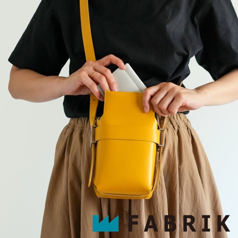 FABRIK 品牌字樣出現在黃色皮革迷你斜背包底部，包包由一位身穿黑色上衣和棕褐色長裙的人士配戴，其雙手正在將白色物品放入包中