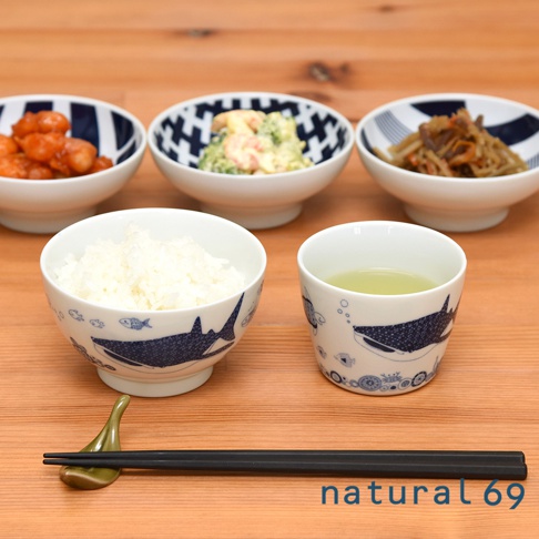 白底藍色鯨鯊與魚圖案的日式飯碗，裝有白飯。右側為同圖案的茶杯，裝有綠茶。後方有三款不同圖案的配菜碗，分別裝有炸雞、沙拉和燉菜。底部有一雙黑色筷子放在綠色筷架上。