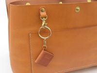 職人手作皮革雙扣兩用側背包，採用天然棕色皮革製成，質感細膩。包包上有金屬扣環和鑰匙圈，鑰匙圈上掛著一個迷你皮革小冊子造型吊飾，增添精緻細節。