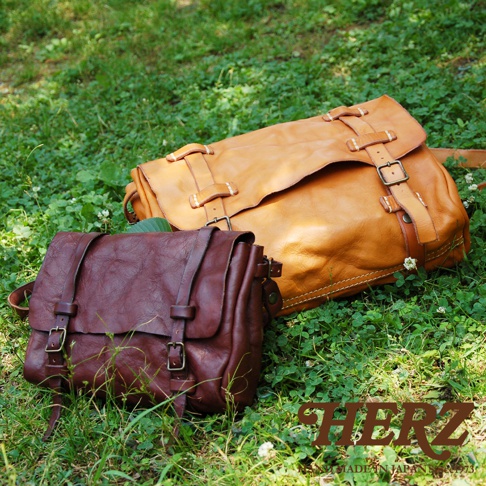 兩款職人手作皮革復古郵差包，一款為深棕色，另一款為淺棕色，均採用水洗加工處理，呈現自然的皮革紋理。兩款包均有翻蓋設計、皮質束帶及金屬釦環，散落在綠色的草地上。