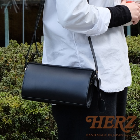 一名穿著白色襯衫與黑色內搭的模特兒，側身背著一個日本HERZ手工製作的黑色極簡風格皮革拉鍊單肩包，包款呈現長筒狀設計，背景為戶外綠色灌木叢。