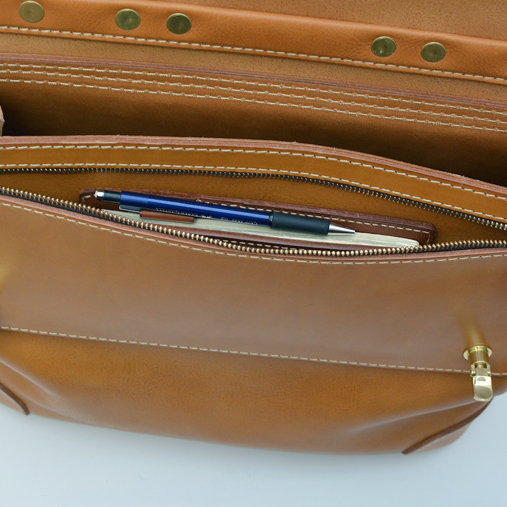 職人手作皮革旅行商務兩用波士頓包，採用光滑棕色皮革，內部有一個拉鍊口袋，內含一支藍色筆和一本筆記本，包身有精緻的縫線和金屬釦。