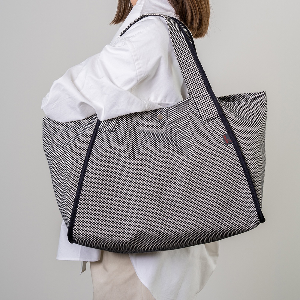 一名女子斜肩背著一個黑色與白色細小格紋圖案的三河木綿刺子織大容量托特包，包包邊緣有深色滾邊。