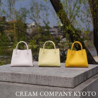 京都職人手作手提托特包，展示淺灰色、淺黃色與亮黃色三款，包身為挺立的梯形，配有對稱的短提把，皮革表面呈現細緻紋理。