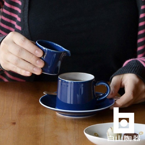 藍色陶瓷糖盅，帶有傾倒口，正被手持著，準備倒入一個藍色陶瓷咖啡杯中，咖啡杯放在同色系的咖啡杯碟上。