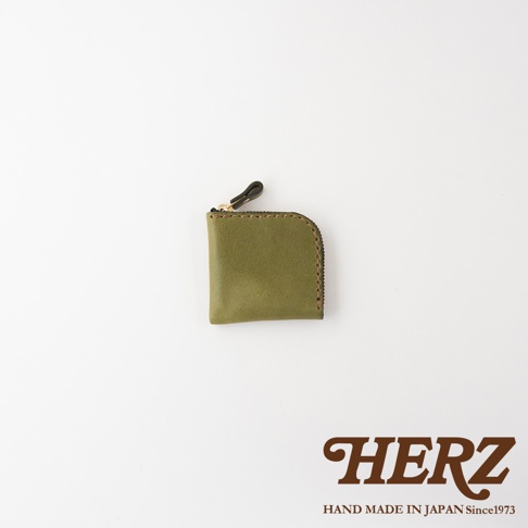 一個由日本 HERZ 職人手作的橄欖綠色皮革零錢包，外觀呈現方形帶有圓弧邊角設計，配有精緻的拉鍊與皮革拉環，右下角印有品牌標誌，整體風格簡約且具有溫潤的皮革質感。
