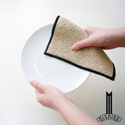 一手拿著一塊米白色、帶有黑色滾邊的棉麻抹布，擦拭著一旁的白色盤子。
