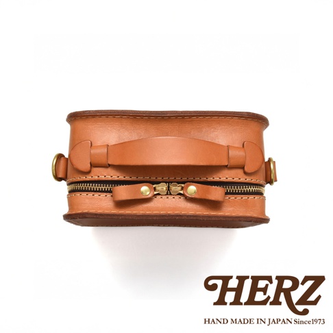HERZ 職人手作皮革小方包，特寫頂部提把與雙拉鍊細節，下方品牌標誌強調日本製造。