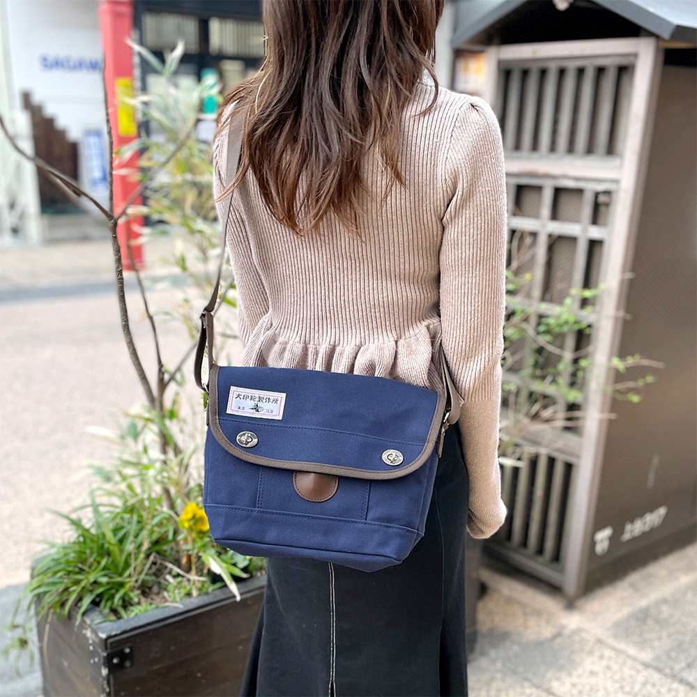 深藍色純棉帆布雙扣郵差小包，配有棕色皮革飾邊及肩帶，包蓋上印有品牌標籤。