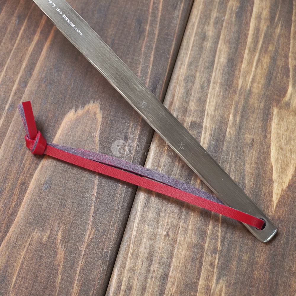 一條金屬色不鏽鋼萬用撈泡勺，勺柄末端繫著紅色彩色皮革與灰色毛氈製成的掛繩，置於木紋背景上。