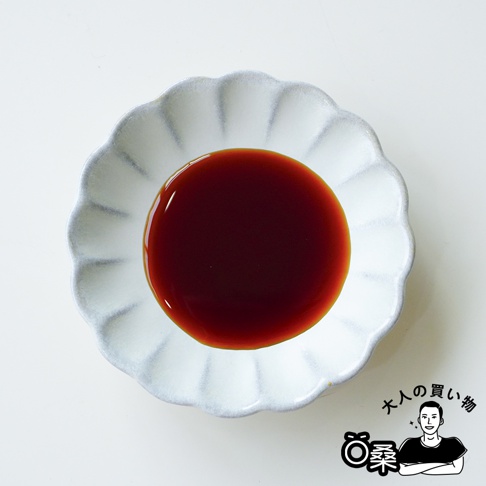 這張俯視照片展示了一只精緻的白色花瓣造型陶瓷碟，碟中盛裝著深褐色、質地清澈的朝紫廣島牡蠣醬油。碟子邊緣呈現微微的灰調暈染質感，與深色的醬油形成鮮明對比。畫面右下角帶有簡約的品牌標誌與人物插圖，整體風格清新雅致。
