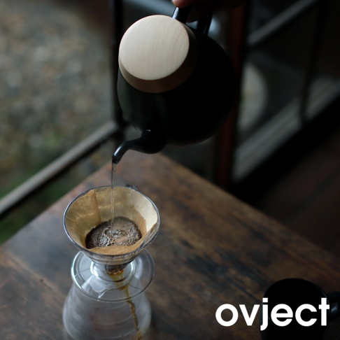 黑色琺瑯手沖壺，搭配木質壺蓋，正從壺嘴傾倒熱水至咖啡濾杯中，濾杯內有咖啡粉與水滴，下方為玻璃分享壺。