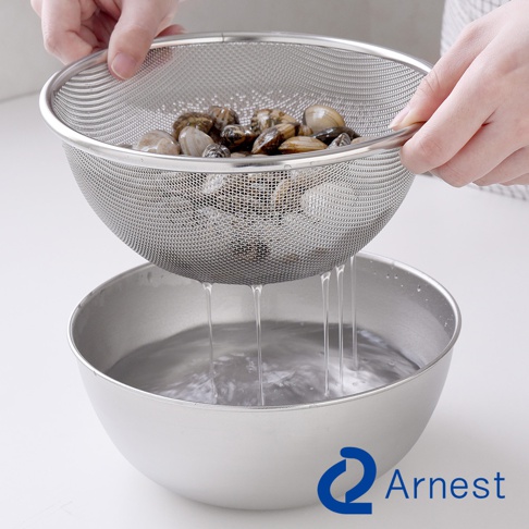 一只手正拿著一個不鏽鋼網篩，篩網裡裝滿了蛤蜊，水正從篩網中滴落到下方一個不鏽鋼盆裡。