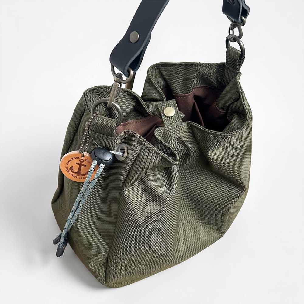 這款橄欖綠色的小型手提包採用防潑水材質，造型簡約俐落，配有黑色皮革提帶與金屬扣環。包身側邊掛有一個印有船錨圖案的神戶clueto品牌圓形皮革吊飾，並設計有束口繩與按扣開合功能，整體風格休閒且具實用性。