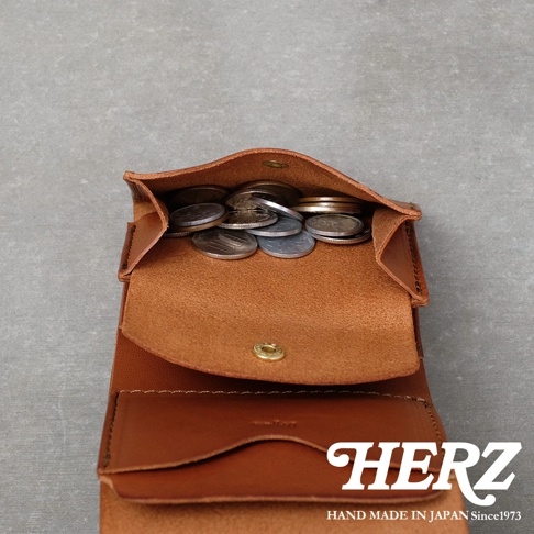 由日本手工皮革品牌HERZ打造的迷你短夾，以溫潤的棕色皮革製成，錢包內部呈現展開狀，上方配有金屬扣環的零錢收納袋中裝滿了日圓硬幣，下方則為卡片夾層設計，整體展現簡約質樸的手作工藝質感。