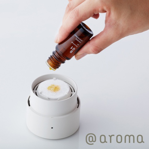手持琥珀色玻璃瓶，將黃色精油滴入白色圓形擴香器內的白色濾芯上，擴香器下方有圓形小孔。