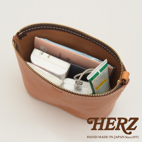 職人手作皮革三角收納包，天然皮革材質，淺棕色，拉鍊開合，包內裝有書籍、充電器、行動電源和牙膏。