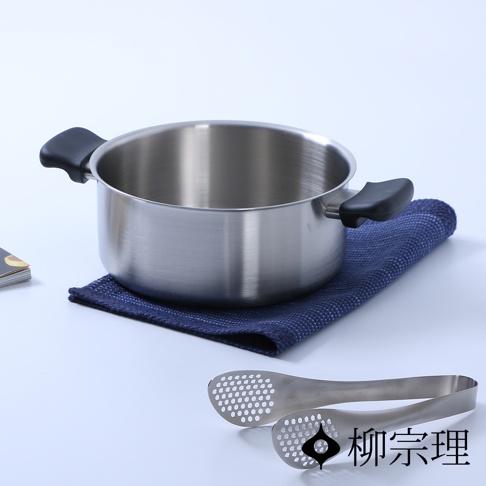柳宗理品牌的不鏽鋼霧面雙耳鍋放置在深藍色編織餐墊上，鍋身呈現簡約的金屬質感，前方擺放一對具有網孔設計的不鏽鋼食物夾，整體畫面為簡潔的商品攝影。