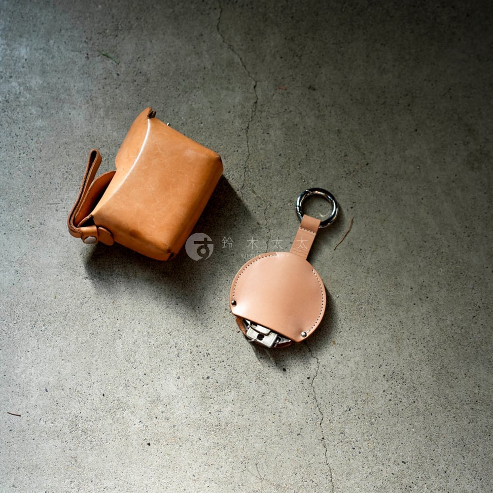 兩個皮革製的日式小物，一個橘棕色的方形小包，配有一條皮帶；另一個是淺棕色的圓形鑰匙圈，上面有金屬環，內部可見鑰匙。