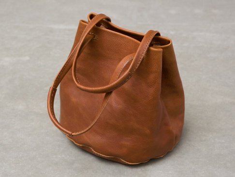 棕褐色皮革抽繩束口托特包，配有兩條手提肩背帶，包身柔軟，整體呈圓潤桶狀。