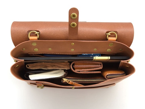 職人手作皮革雙扣兩用側背包，呈現原皮色澤，內部整齊收納了筆記本、皮夾、筆和其他小物，展現皮革質感與實用性。