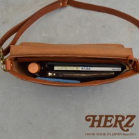 HERZ 品牌標誌，下方為 HAND MADE IN JAPAN Since1973 字樣，整體為皮革隨身斜背包的內部視角，包內裝有書籍、小皮夾、藍色布料及一瓶飲料。