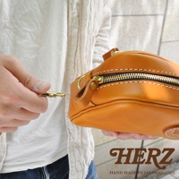 職人手作的橘棕色皮革萬用手提包，特寫拉鍊與可拆式扣環，展示皮革材質的紋理與細節。