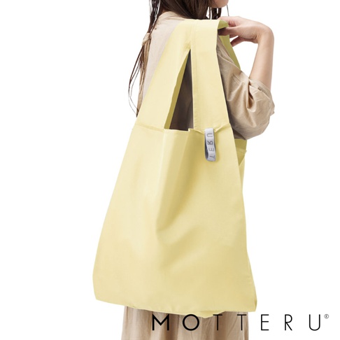 一位女性左肩上搭著一個淺黃色的大容量摺疊環保隨行袋，袋子為尼龍材質，附有同色系的提把，側面有一個灰色小標籤。