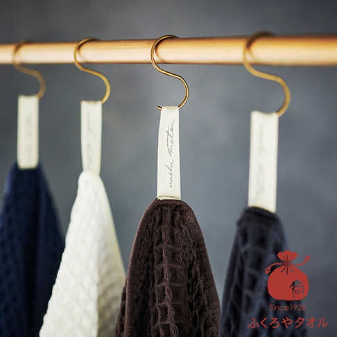 四條掛在銅桿上的毛巾，分別為深藍色、白色、咖啡色和深藍色，每條毛巾都附有標籤，其中一條標籤上寫著 washi maton