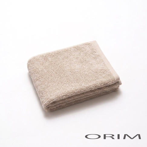 一張放置於白色背景上的 ORIM 品牌淺棕色純棉毛巾，毛巾質地蓬鬆柔軟，以整齊的長方形摺疊呈現，右下角印有深灰色 ORIM 品牌字樣，整體展現簡約質感。
