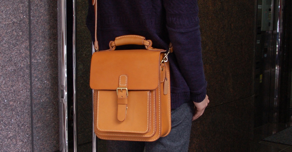 咖棕色皮革單肩兩用小方包，設有翻蓋、皮帶扣和手提把，搭配米白色縫線，展現職人手作質感。