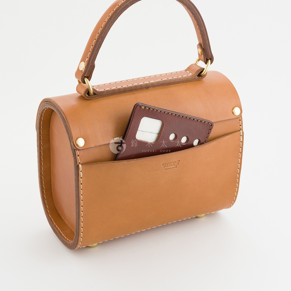 職人手作皮革復古杜勒斯手提包，採用咖啡色皮革製成，設有外部收納袋，可放置卡片或名片，包身線條圓潤，搭配金色旋鈕扣與提把，展現經典復古風格。