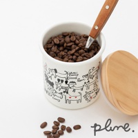 白色琺瑯儲物罐，外觀印有黑色線條描繪的各式狗狗圖案，罐內裝有咖啡豆，一把木柄金屬勺插入罐中。旁邊擺放著一個木質圓形蓋子，以及幾顆散落的咖啡豆。