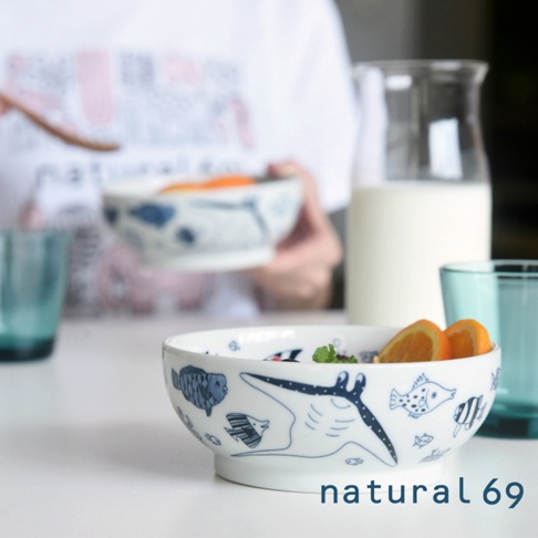 白色陶瓷小丼碗，碗身繪有藍色海洋生物圖案，包括魚和魟魚。碗內盛有麥片、香蕉片和橘子片。背景有兩個藍色玻璃杯和一個裝有牛奶的玻璃瓶。