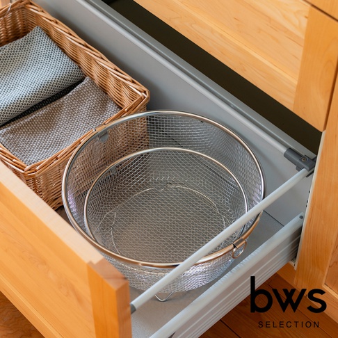 一個淺型的圓形不鏽鋼網狀瀝水籃，籃身佈滿細密網孔，邊緣有金屬環。旁邊有一個藤編織的籃子，裡面放著兩塊淺灰色的布。