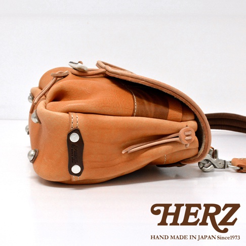 淺棕色皮革製成的郵差包，側面有鹿角狀的扣環設計，包身有金屬铆釘裝飾，搭配深棕色皮革背帶，整體呈現職人手作質感。