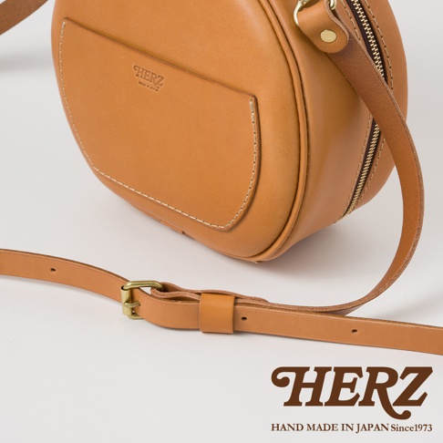 日本HERZ手作皮革圓形包，咖啡色，帶有金屬扣環和可調式皮革背帶，下方壓印有HERZ品牌標誌。