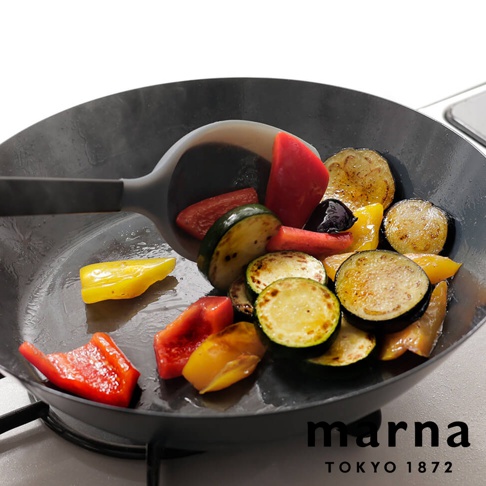 marna TOKYO 1872 品牌標誌，以及一個黑色平底鍋，鍋內盛有炒熟的蔬菜，包含櫛瓜、茄子、紅椒和黃椒，一把灰色矽膠料理匙正從鍋中撈起部分蔬菜