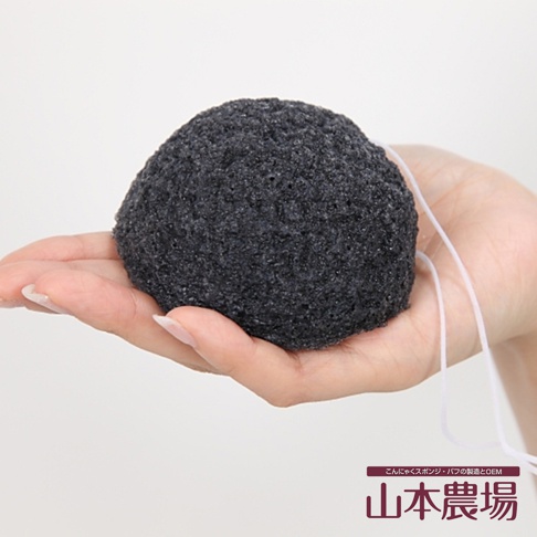 手持一顆深灰色、質地粗糙的蒟蒻洗臉海綿，海綿呈圓球狀，表面有許多細小孔洞，一條細白色掛繩從海綿後方延伸而出。