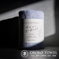 一條淺藍色的有機棉浴巾，質地柔軟，捲起後標籤上印有日文和英文說明「やさしいオーガニック バス タオル」、「SIZE: 約60CM×120CM」、「Made in Japan / cotton 100%」及「OBORO TOWEL」字樣。