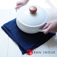 雙手捧著白色鍋子，鍋蓋上有木製把手，鍋子下方墊著深藍色棉麻布料。