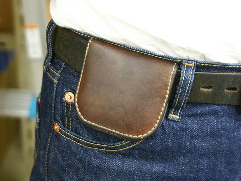 職人手作皮革馬蹄造型零錢包，深棕色皮革，縫線細緻，搭配牛仔褲與皮帶，展現復古風格。