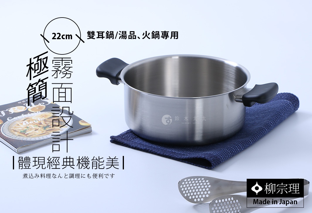 圖片中呈現柳宗理22公分霧面不鏽鋼雙耳鍋，鍋身具有質感霧面設計，放置於深藍色餐巾布上。左側有一本烹飪食譜，右下方配有不鏽鋼濾網夾，整體風格簡約雅致，展示日本製造的經典機能美學。
